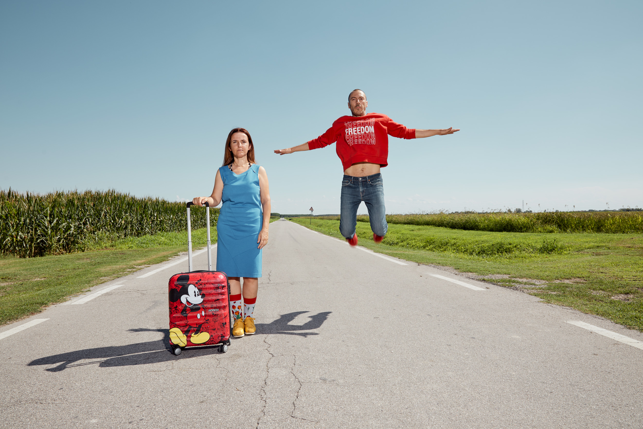 Due persone, un uomo e una donna, stanno in mezzo a una strada di campagna. L'uomo salta, la donna tiene in mano una valigia.