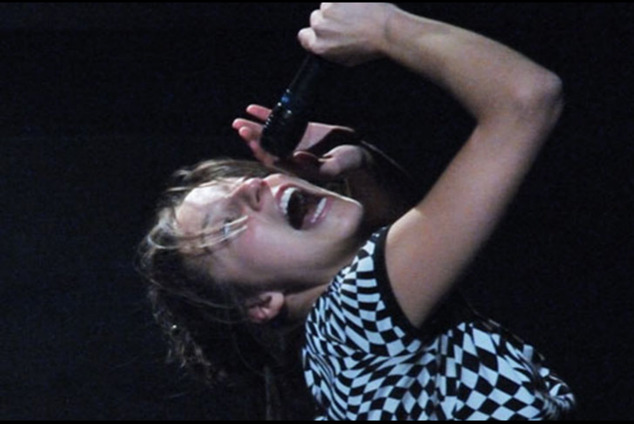 Una ragazza urla al cielo, reggendo con le mani un microfono.