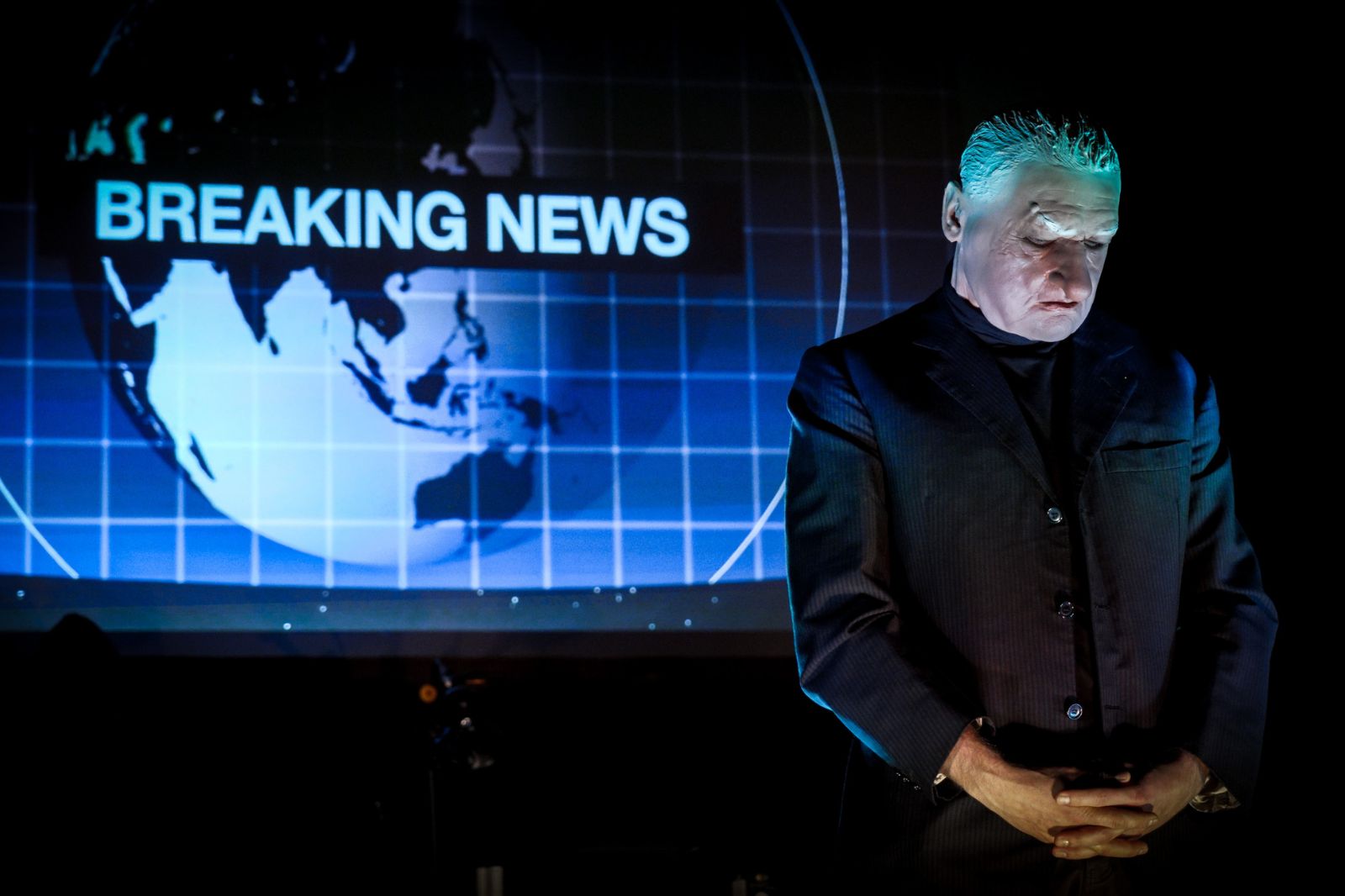 In foto, un uomo indossa una maschera dando le spalle a uno schermo dove viene proiettato il notiziario