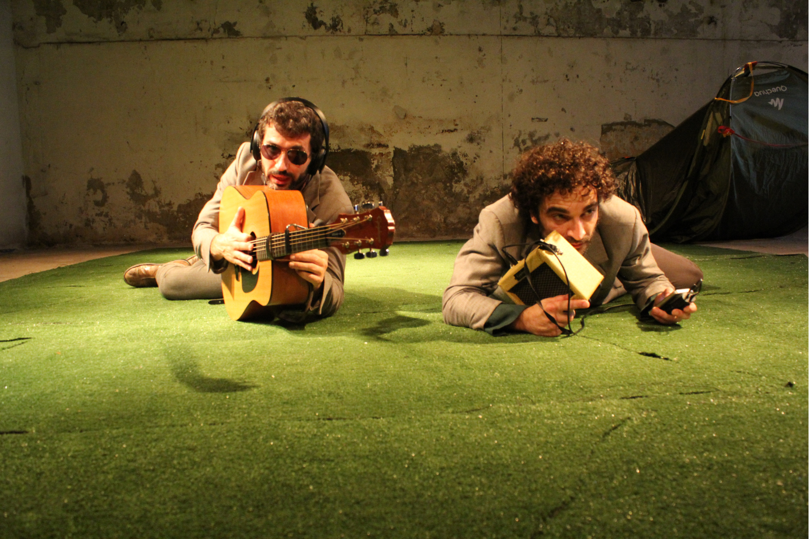 In foto, due uomini sdratiati a terra, uno imbraccia una chitarra, l'altro tiene in mano una radio